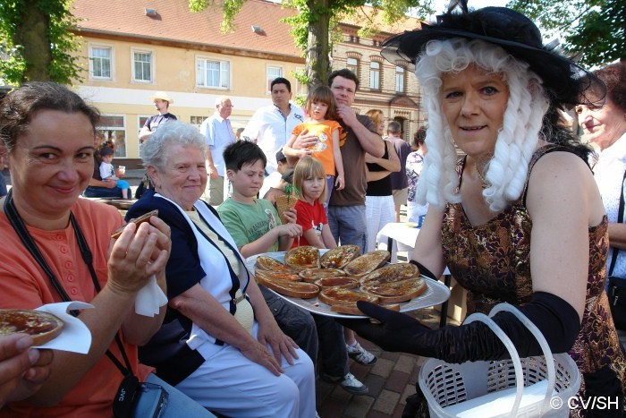 Bild: Anlässlich des 55-jährigen Bestehens des Zörbiger Saftjungen auf dem Zörbiger Marktplatz, gab es einen geschichtlichen Rückblick zur Entstehung der Stein-Skulptur auf dem Springbrunnen. Mit Zörbiger Rübensaft bestrichene Brote verteielten die Stadtdamen an die anwesenden Gäste.